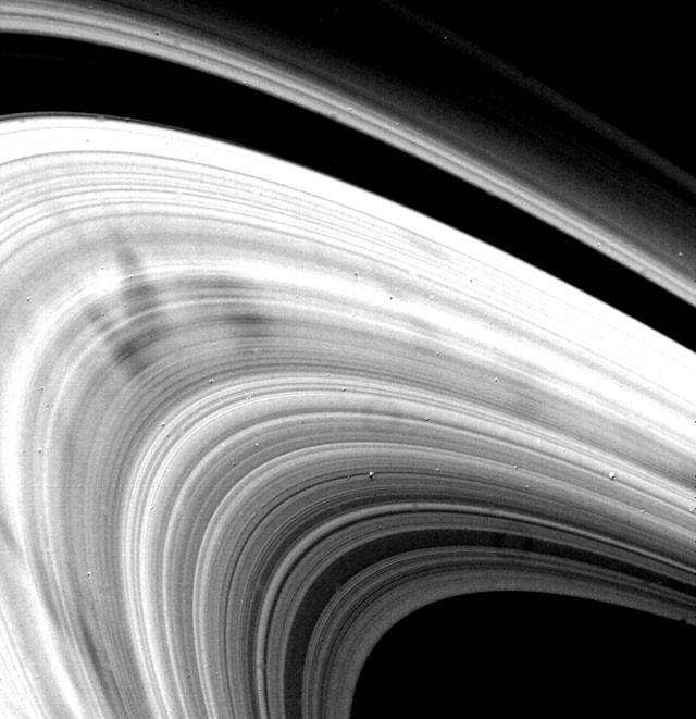 Aproximação da Voyager 1 dos anéis de Saturno que possui várias luas riscando sua estrutura. 